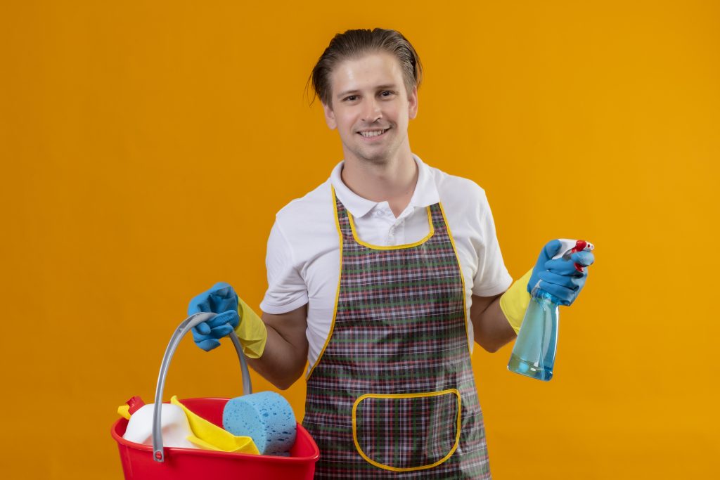 young hansdome man wearing apron rubber gloves holding bucket with cleaning tools